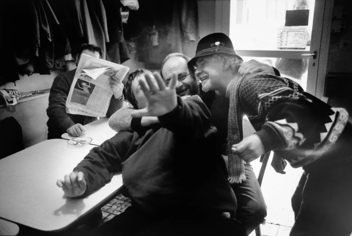 Men around a long table in a casual indoor space; one man waves at the camera while others smile, and another reads a newspaper.