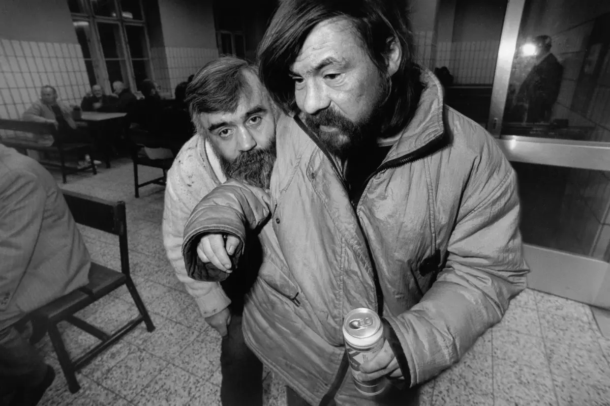 Two bearded individuals in jackets lean close in a tiled indoor space; one holds a can while others sit at tables in the background.