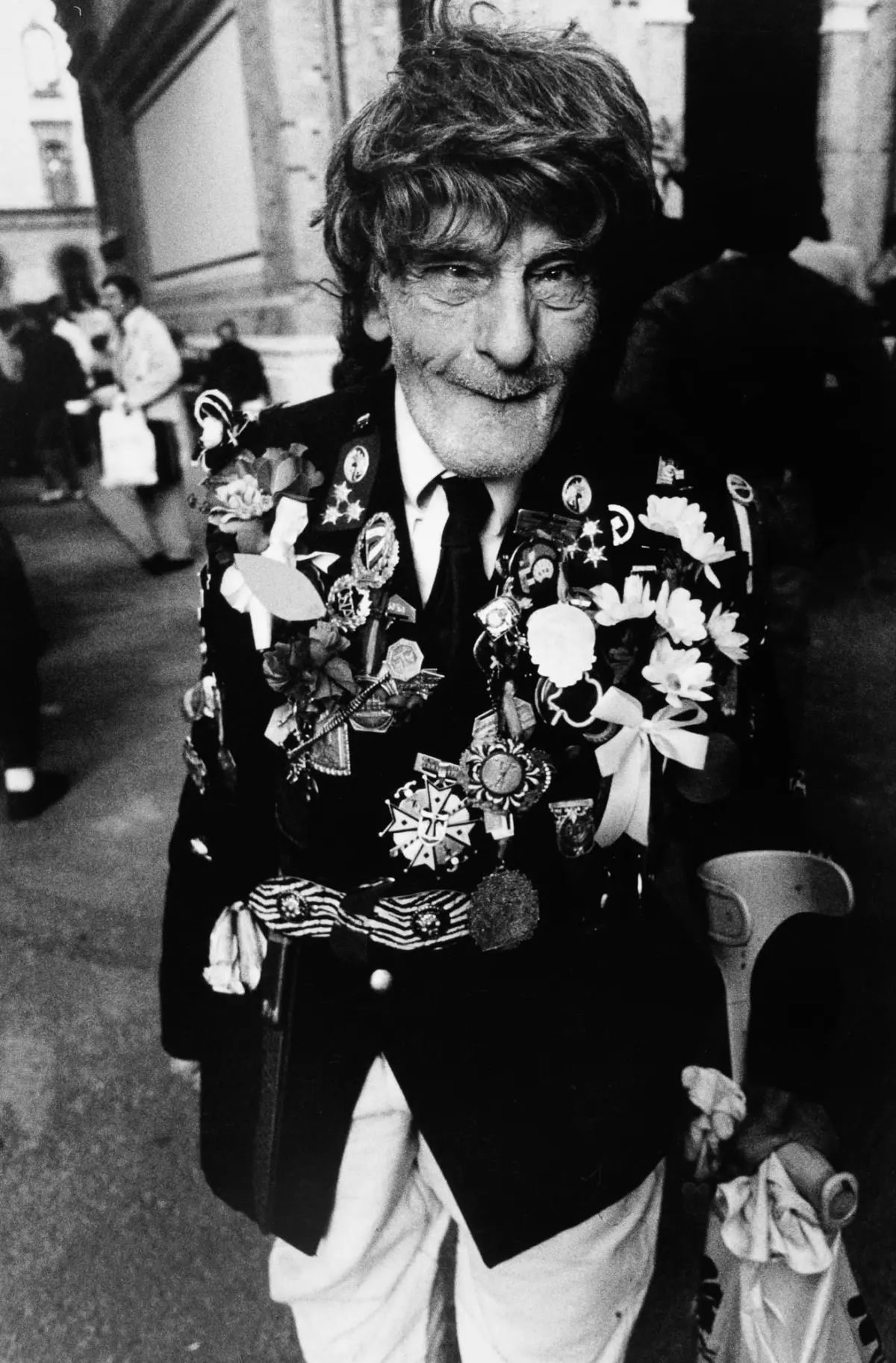 Older man wearing a medal-covered blazer walks through a street parade, smiling.