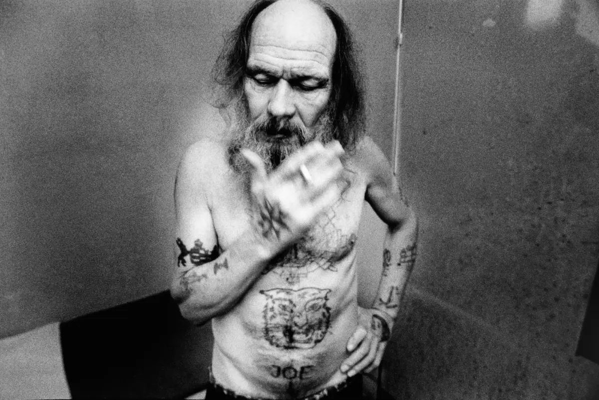 Adult person with long hair and beard, tattooed chest and arms, smoking a cigarette indoors.