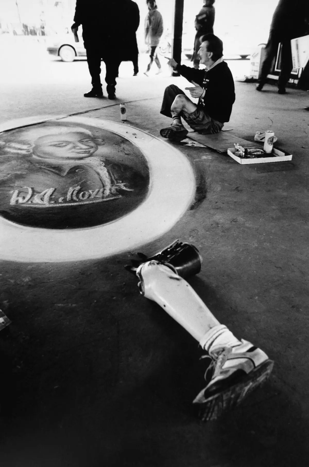 Street chalk artist sitting on cardboard, drawing a portrait in a circular mural on the sidewalk; art supplies nearby, pedestrians in background.