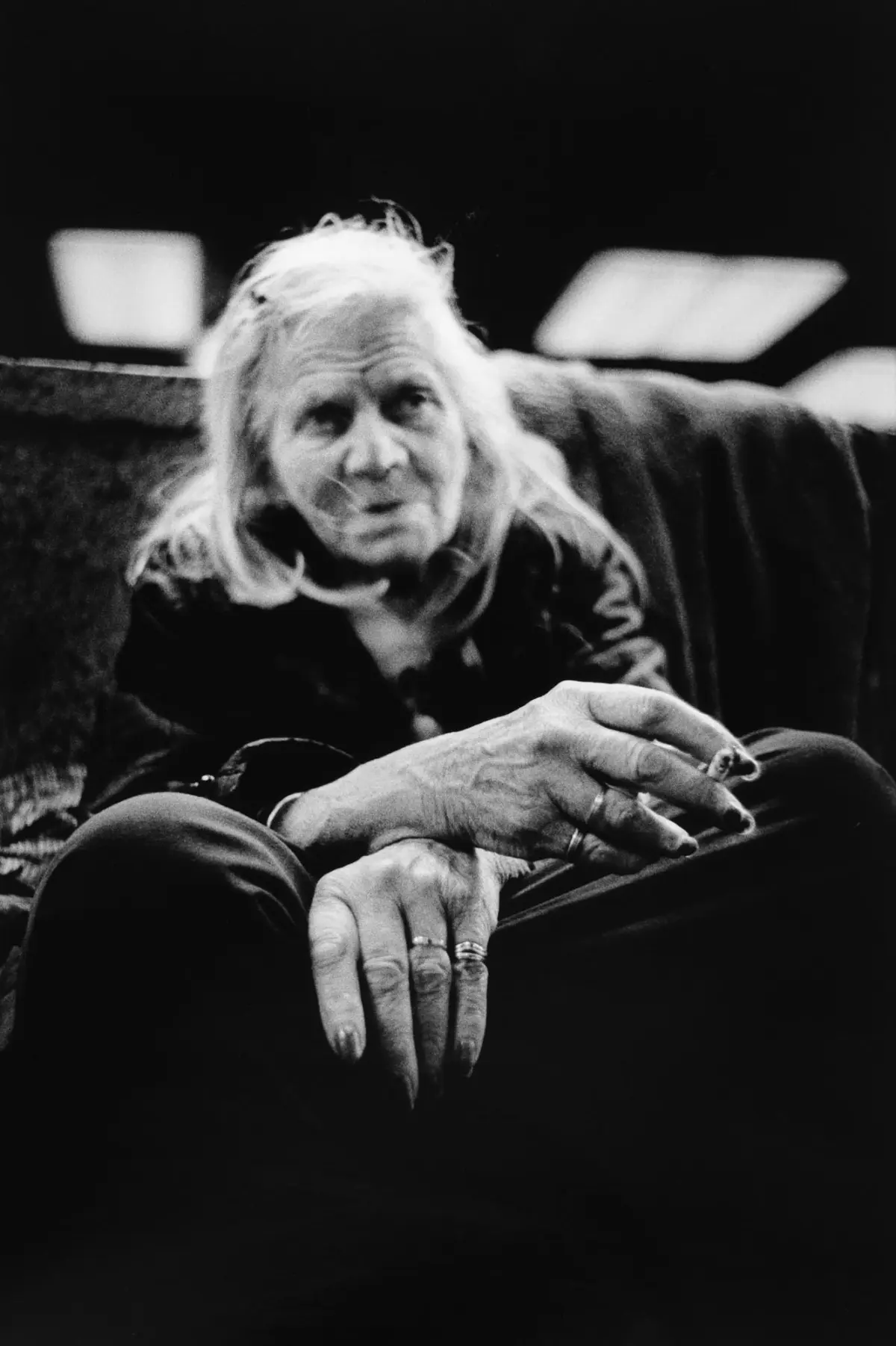 Elderly person with long white hair sits, leaning forward, hands folded on lap, rings visible, in a dim indoor setting.