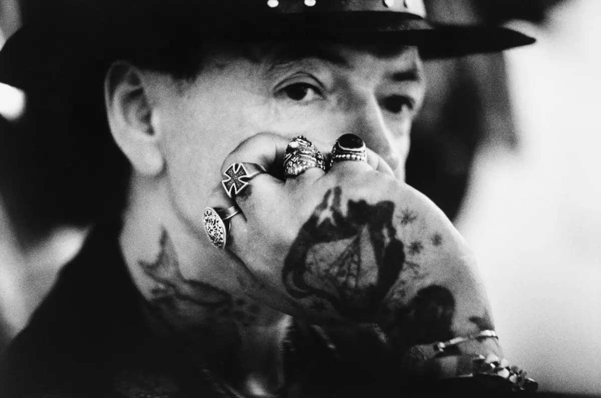 Person with tattooed arm and multiple rings touching lips in a close-up black-and-white portrait, wearing a hat.