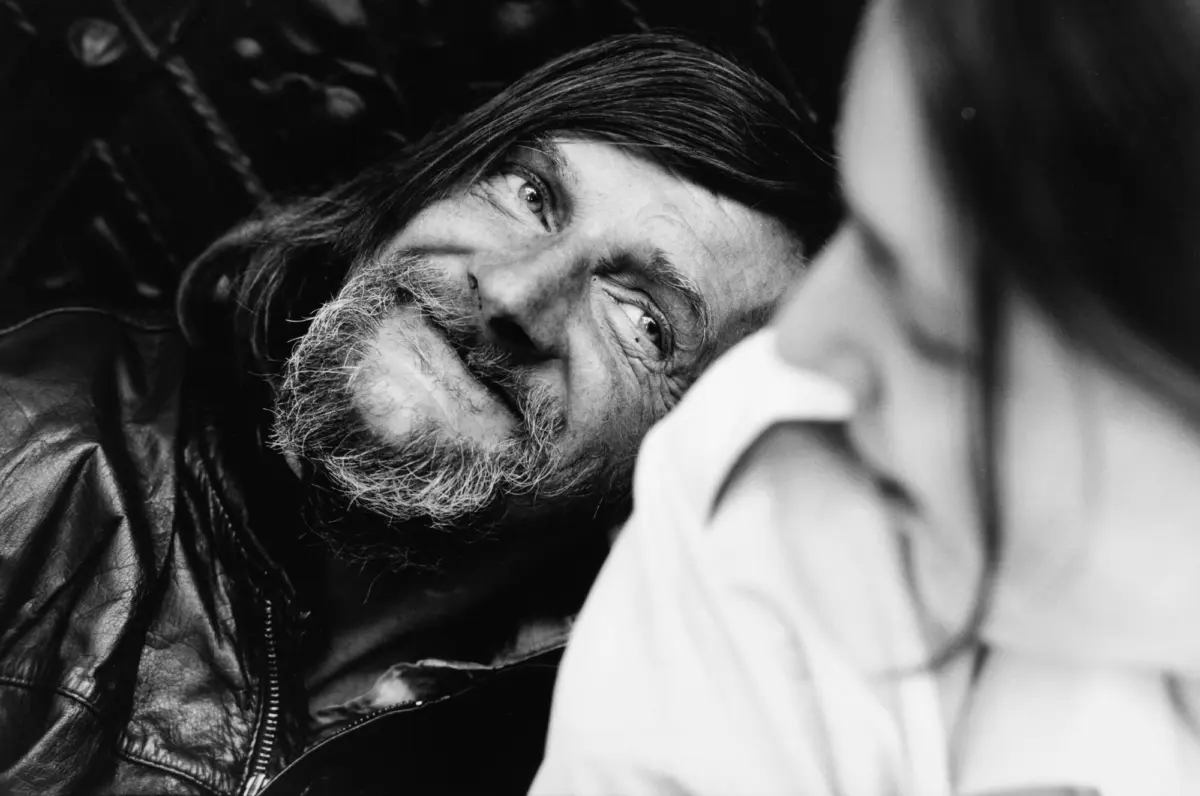 Close-up of an older man with a gray beard and long hair, smiling, wearing a leather jacket.