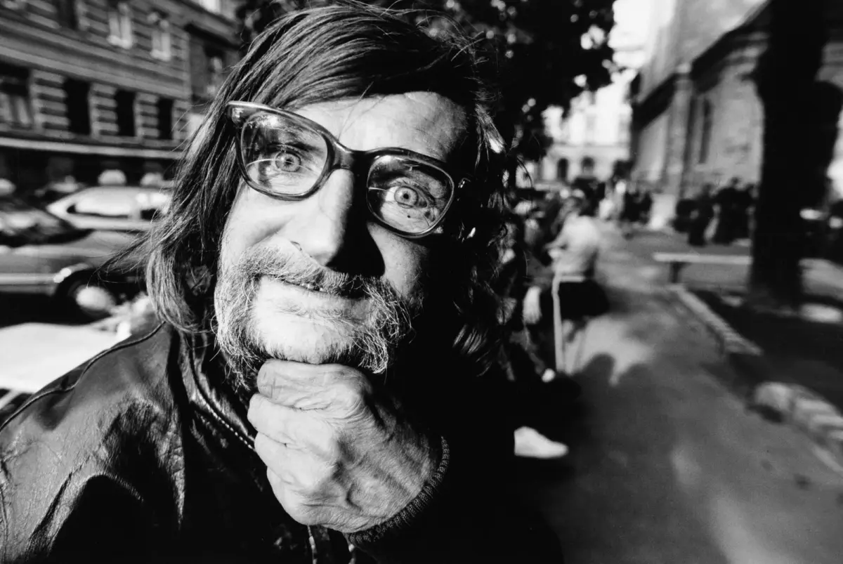 Portrait of a man with long hair and beard wearing thick glasses, smiling with his hand under his chin, on a city street.