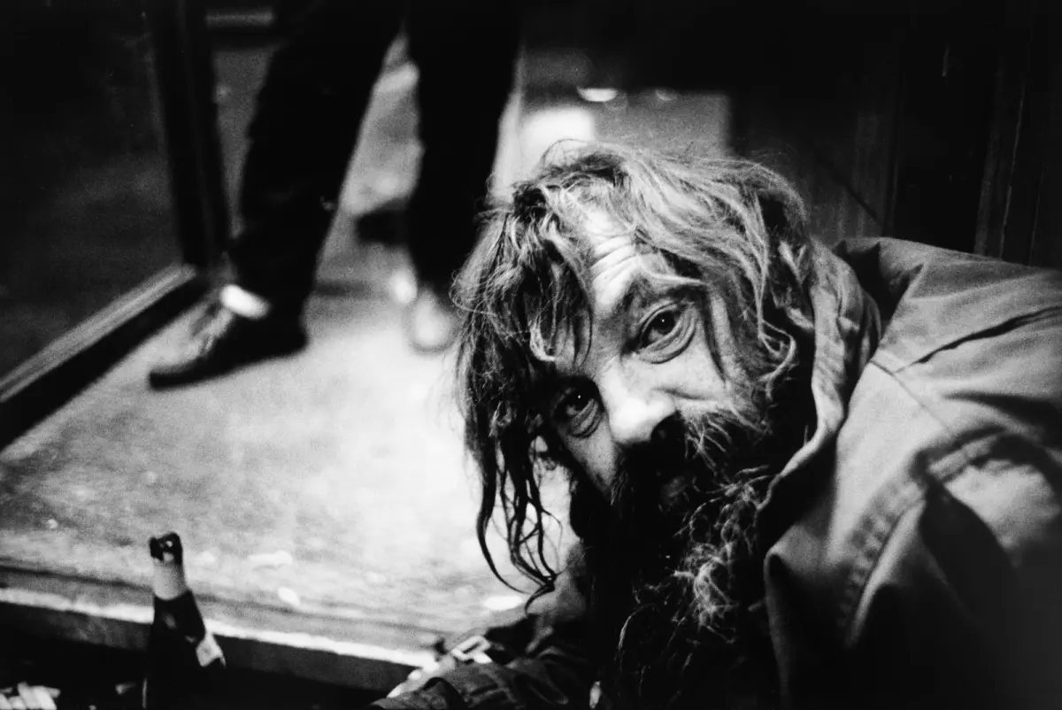 Disheveled person with long hair lying on the floor, looking at the camera; blurred figure in doorway behind.