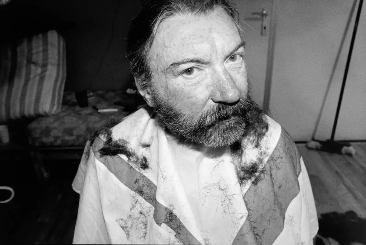 Adult male with thick beard and long hair, looking to the right, wearing a diagonally striped shirt, in a cluttered indoor room.