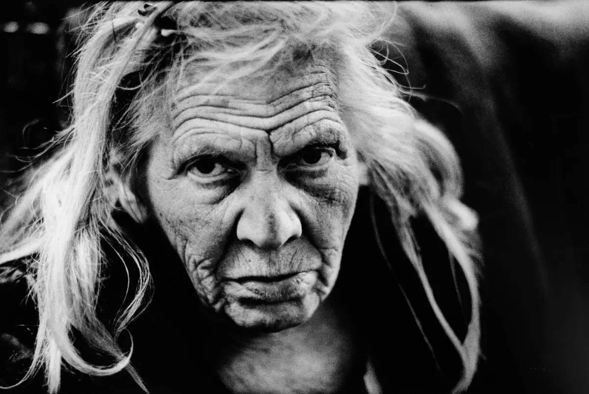 Close-up portrait of an elderly person with long gray hair, deep wrinkles, and a somber gaze against a dark background.