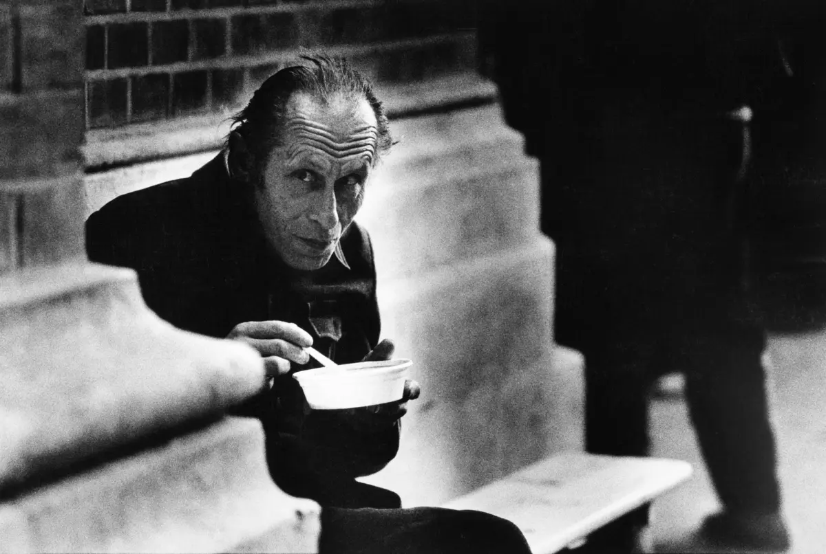 Older person with slicked-back hair sits on stone steps outdoors, eating from a white bowl with a spoon, glancing at the camera.