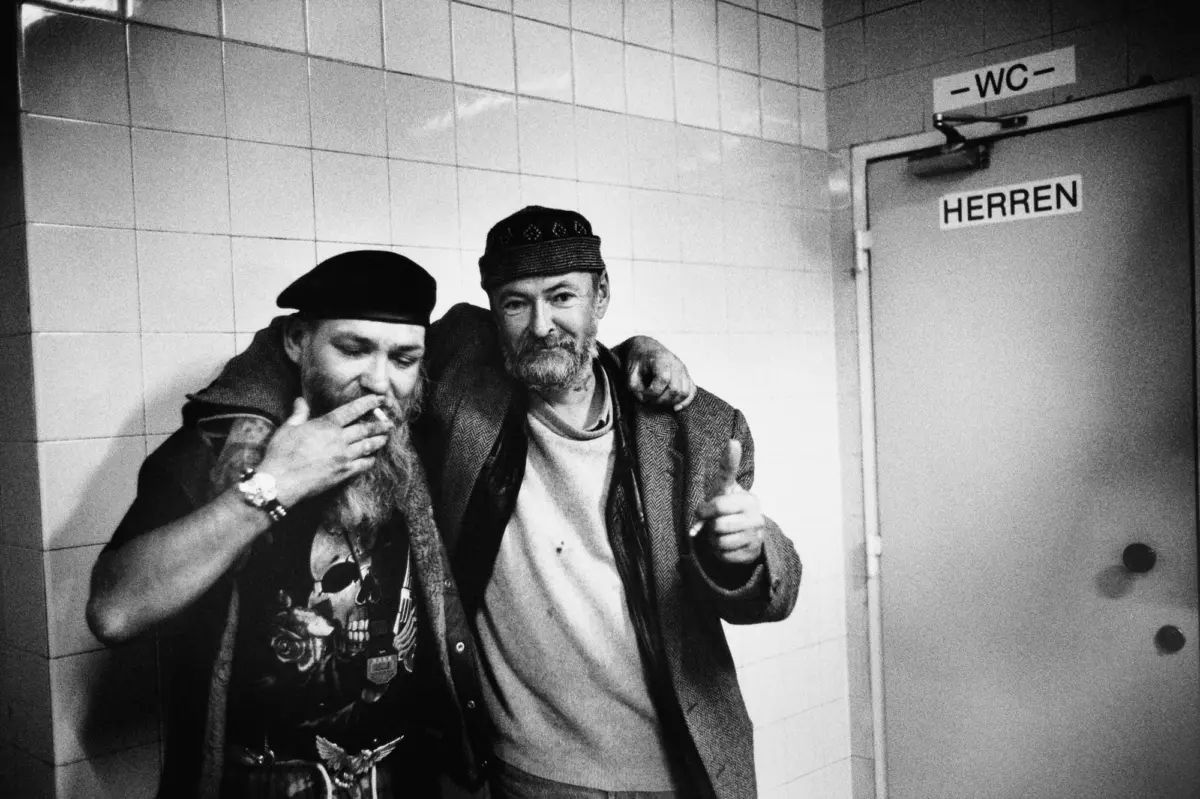 Two adults with arms around each other pose in a tiled hallway by a door labeled HERREN with a -WC- sign above; one smokes.