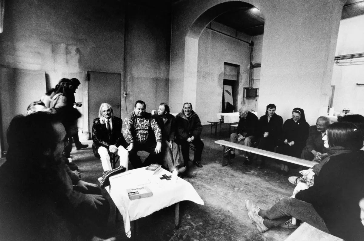 Group of adults seated in a circle in an industrial room, engaged in conversation; table with books in the foreground.