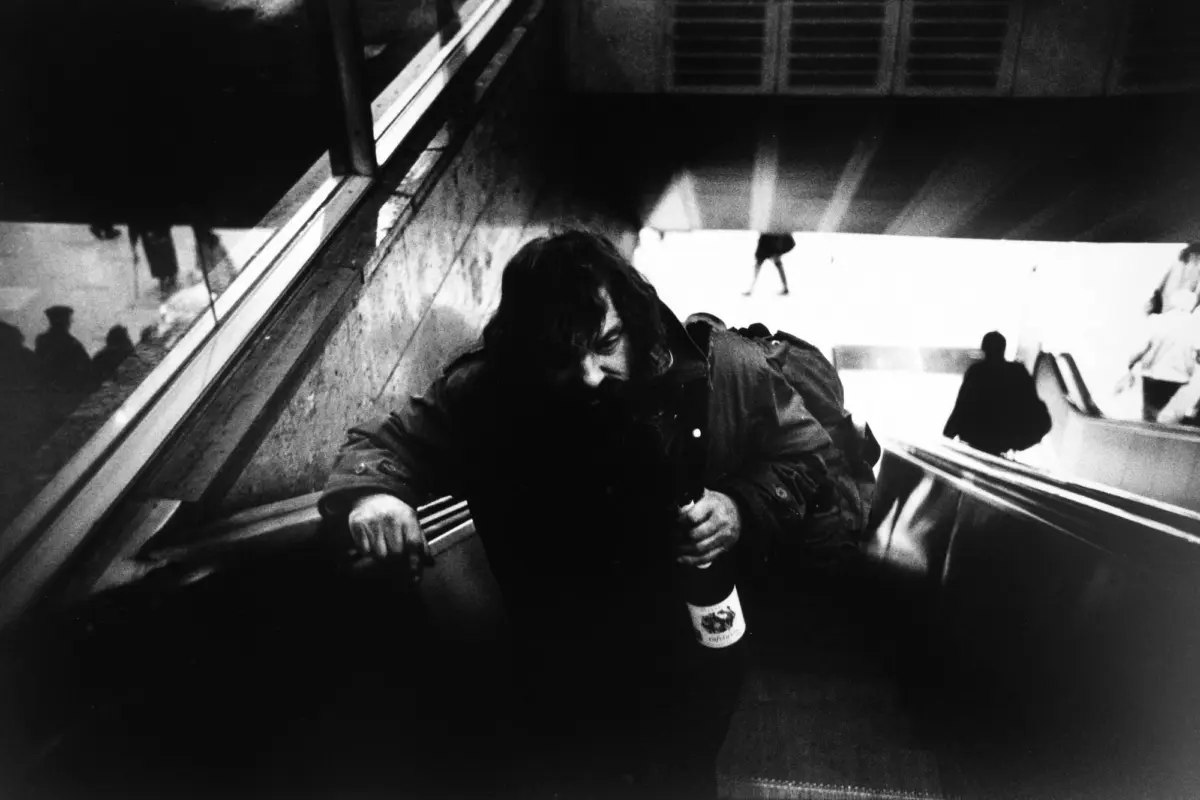 Person on a subway escalator, leaning forward and gripping the rail while holding a bottle.