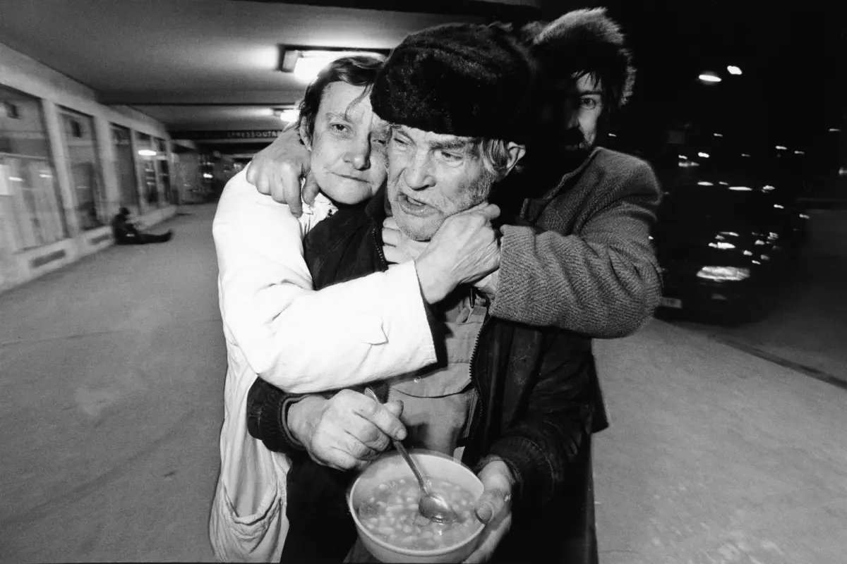 Two men embrace on a nighttime subway platform; one wearing a white lab coat holds a bucket of food, the other wearing a fur hat.