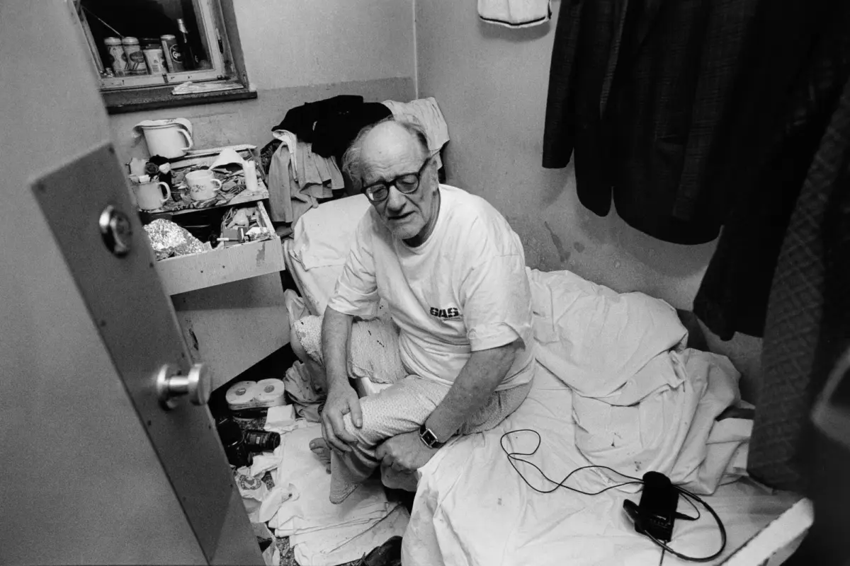 Older adult with glasses sits on a rumpled bed in a cramped, cluttered room, surrounded by clothing, cups, and scattered items.