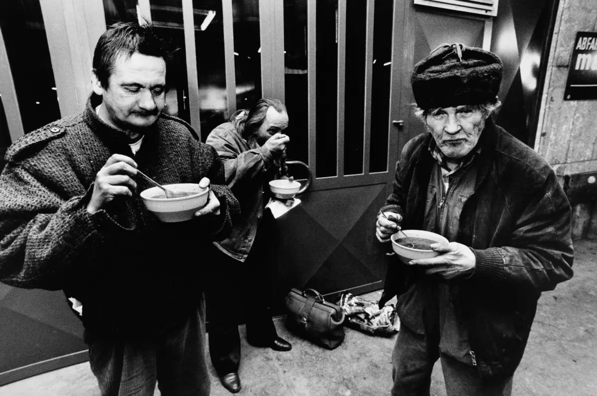 Three adults eating from bowls outdoors; left adult in thick knitted sweater, middle adult covers mouth, right adult in fur hat and jacket.