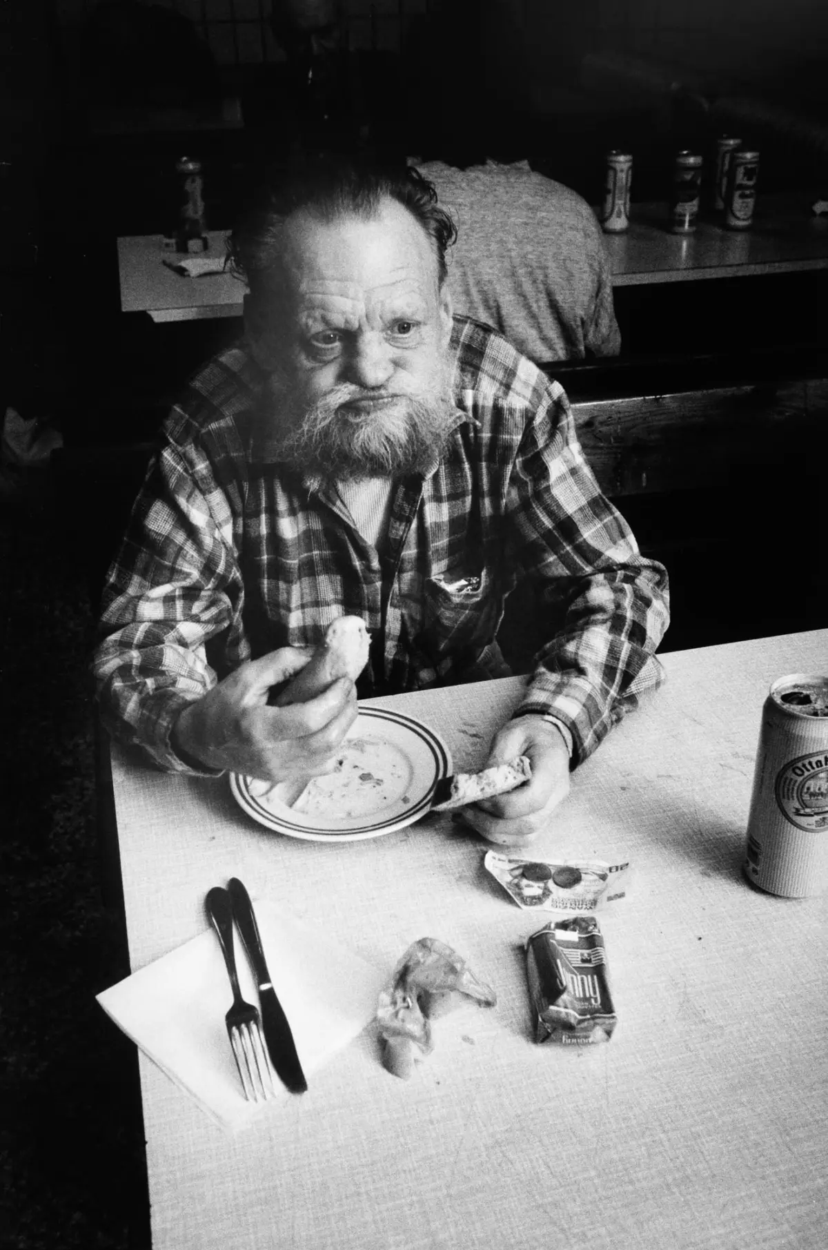 Older man with a beard in a plaid shirt eats a sandwich at a table in a casual dining area.