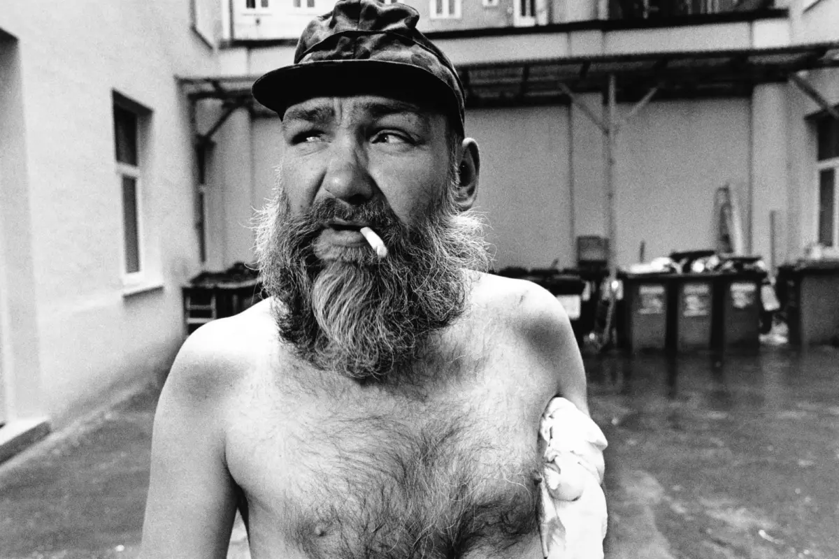 Bearded man wearing camouflage cap, shirtless, smoking a cigarette, in an outdoor alley with dumpsters.