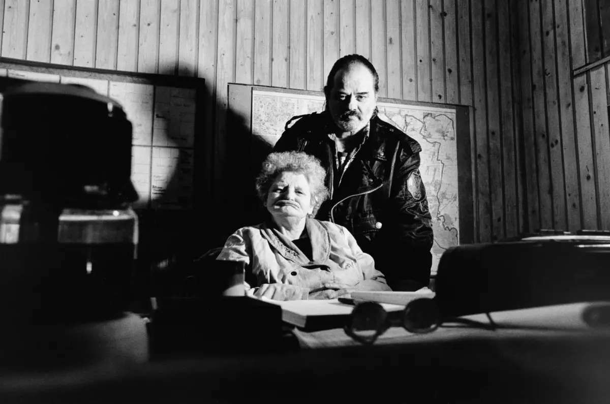 Elderly woman seated at a desk with papers; man in a leather jacket stands behind her in a wood-paneled room with maps.