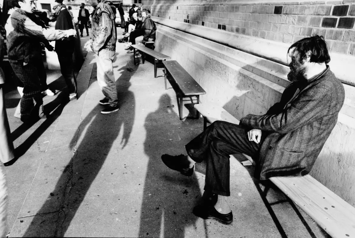 Bearded man in a coat sits on a bench along a brick wall, looking downward; urban crowd nearby.