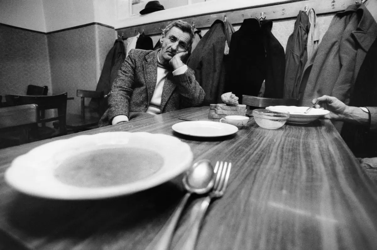 Older man in a tweed blazer rests his head on his hand at a long wooden table, coats hanging on the wall behind in a cafe.