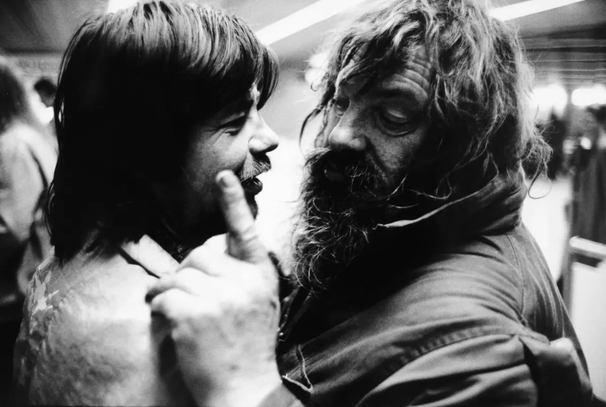 Two adults hug closely in a subway station; one has short hair and mustache, the other long hair and beard.