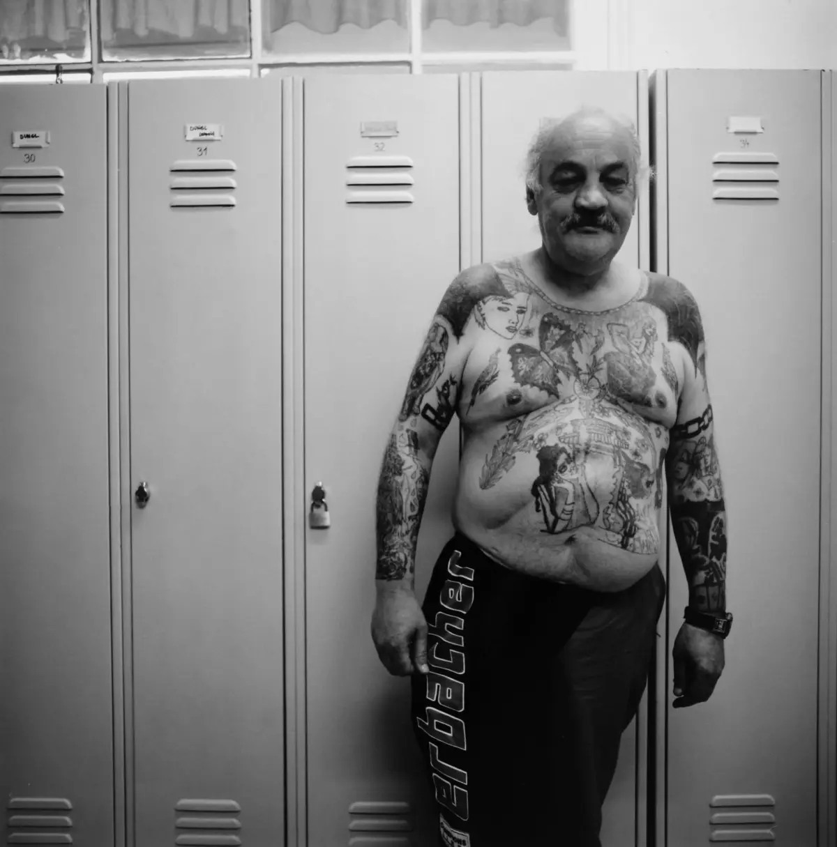 Tattooed, middle-aged man stands shirtless in a locker room, eyes closed, in front of a row of lockers.