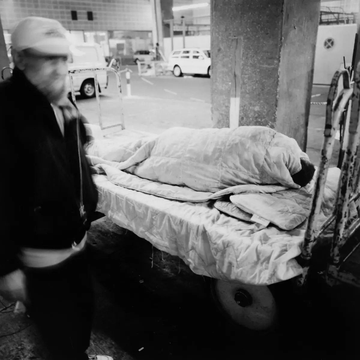 Blurred person wearing a cap beside a pile of quilts on a cart in an urban street.