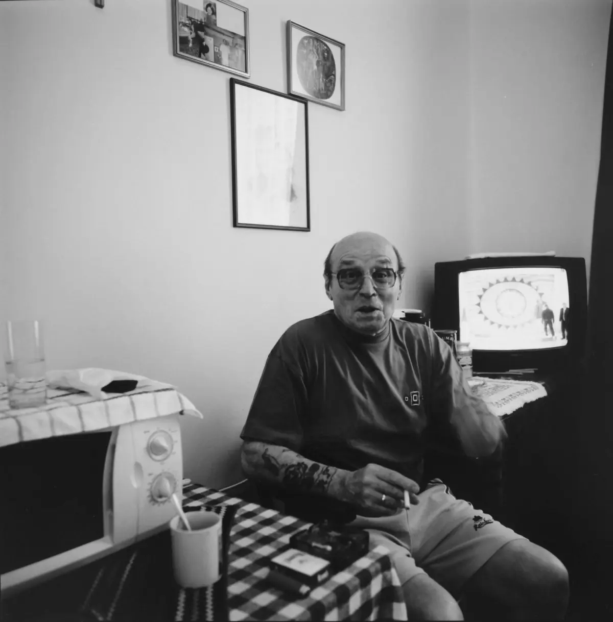 Elderly man with glasses sits at a checkered table, smoking a cigarette, in a room with framed pictures and a retro television.