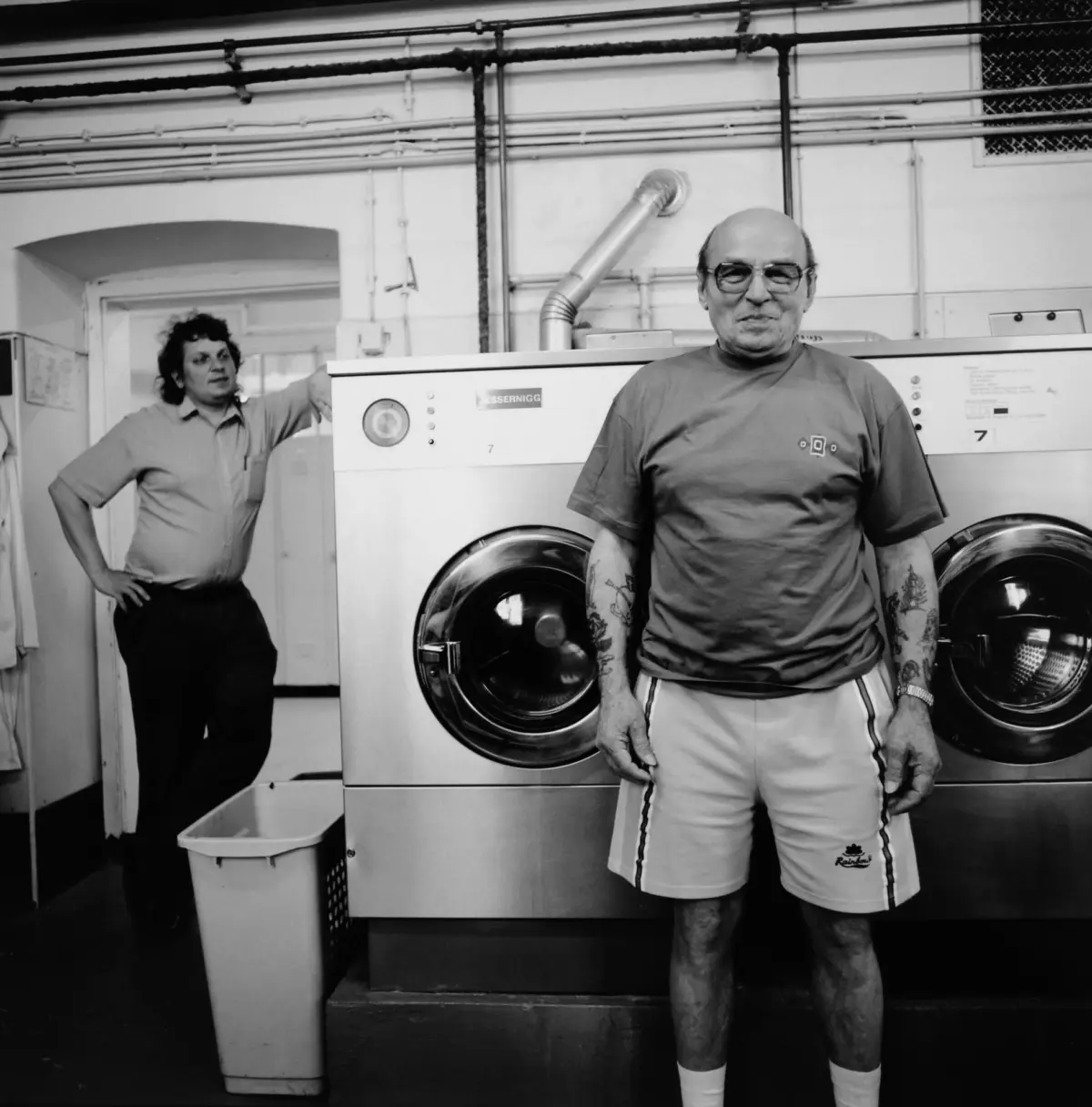 Older man with tattoos and glasses standing in front of a front-loading washing machine in a laundromat.