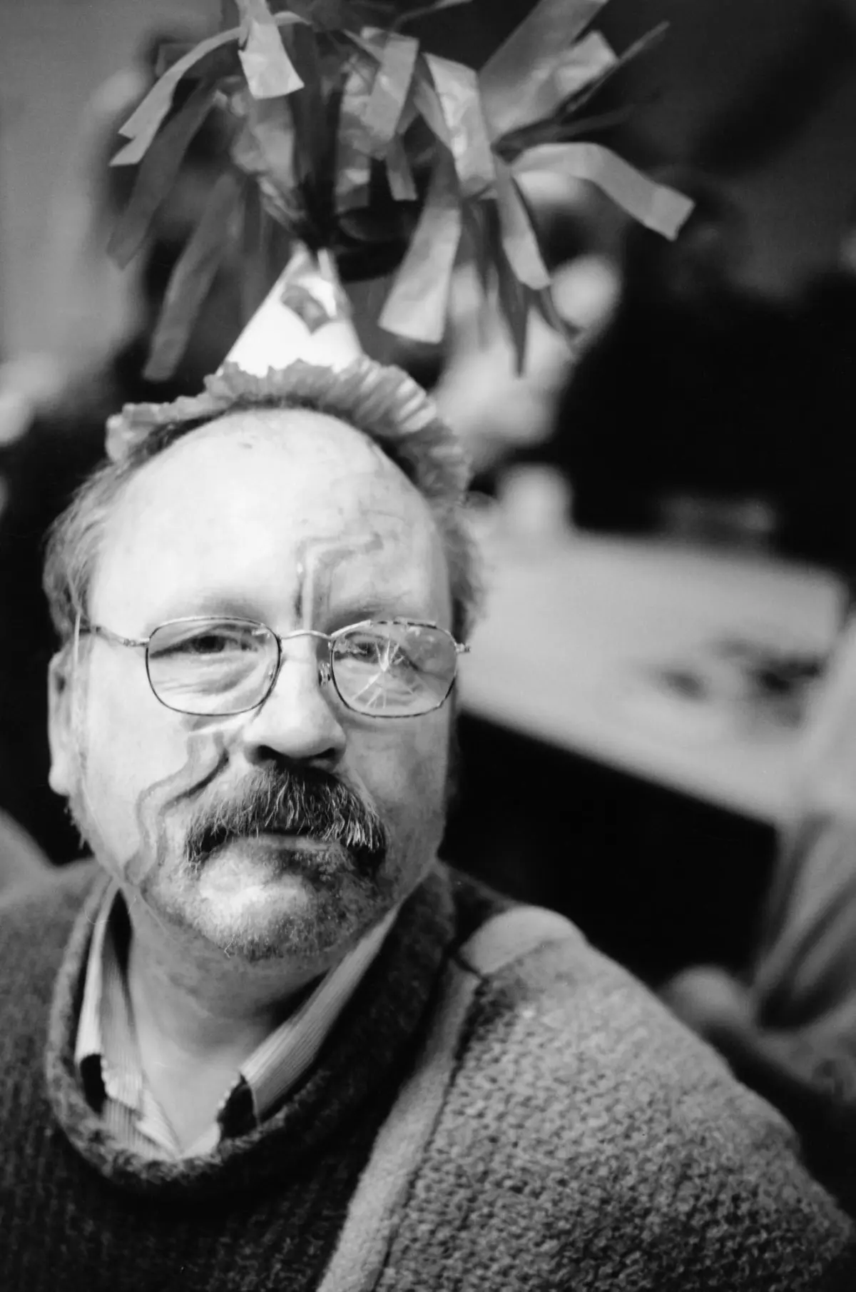 Adult man with glasses wearing a paper party hat at a celebration, confetti streamers overhead.