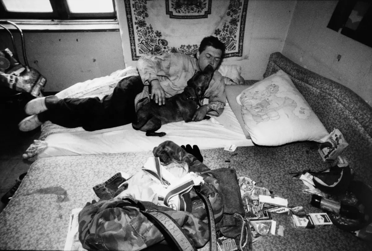 Person lying on a bed as a dog licks their face in a cluttered bedroom.