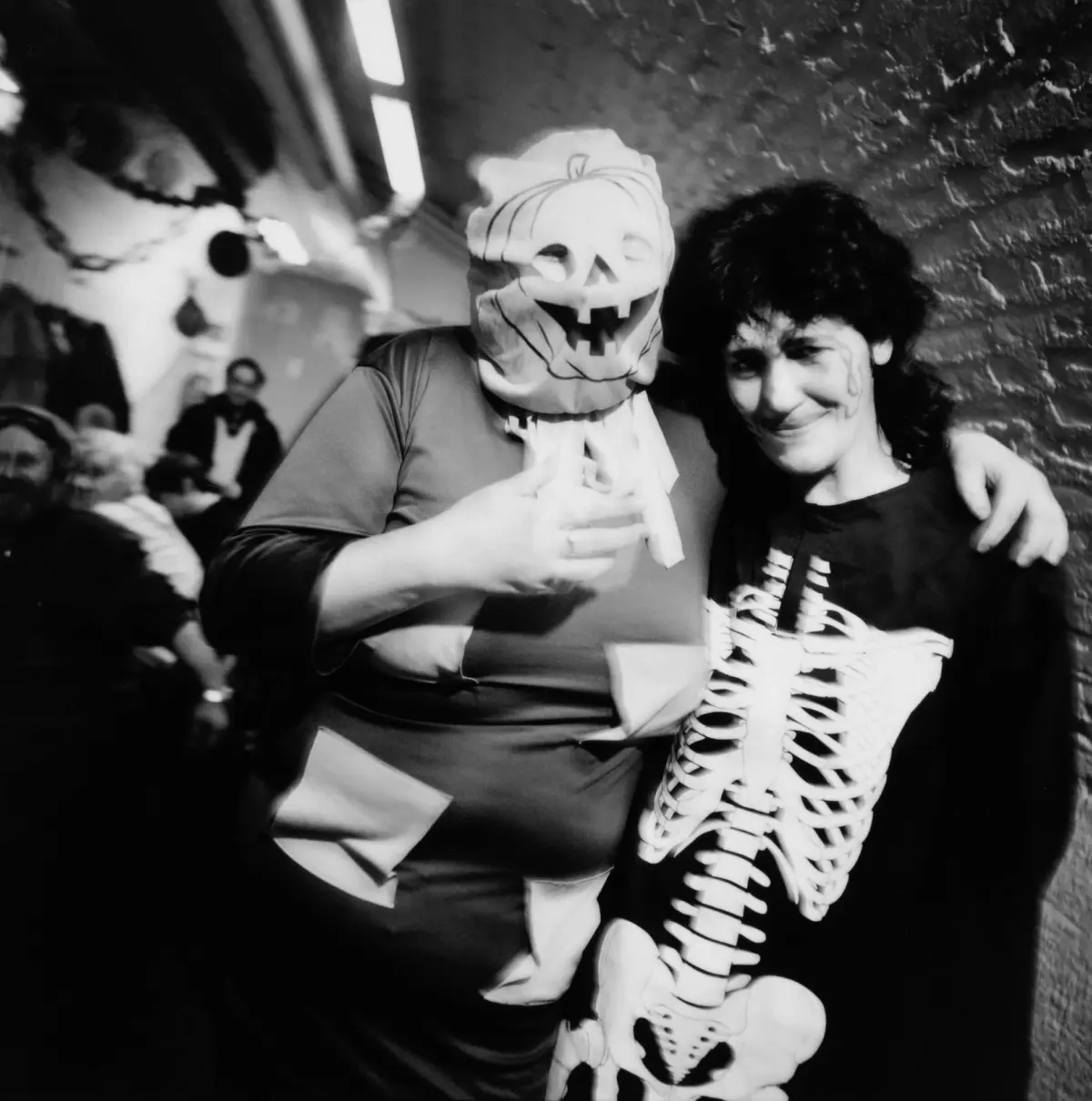Two people in Halloween costumes posing together; one with a jack-o'-lantern mask, the other in a skeleton-themed outfit, in a dim brick corridor.