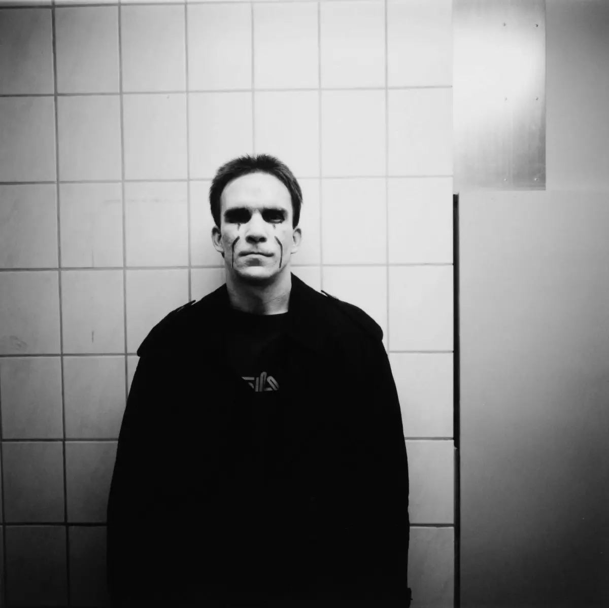 Black-and-white photo of a person with facial paint markings standing against a tiled wall, facing the camera.