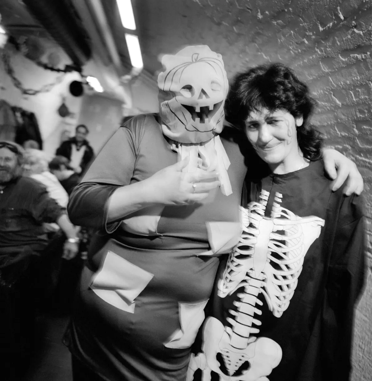 Two people in Halloween costumes pose together; one wearing a pumpkin mask, the other with skeleton makeup, against a brick wall.