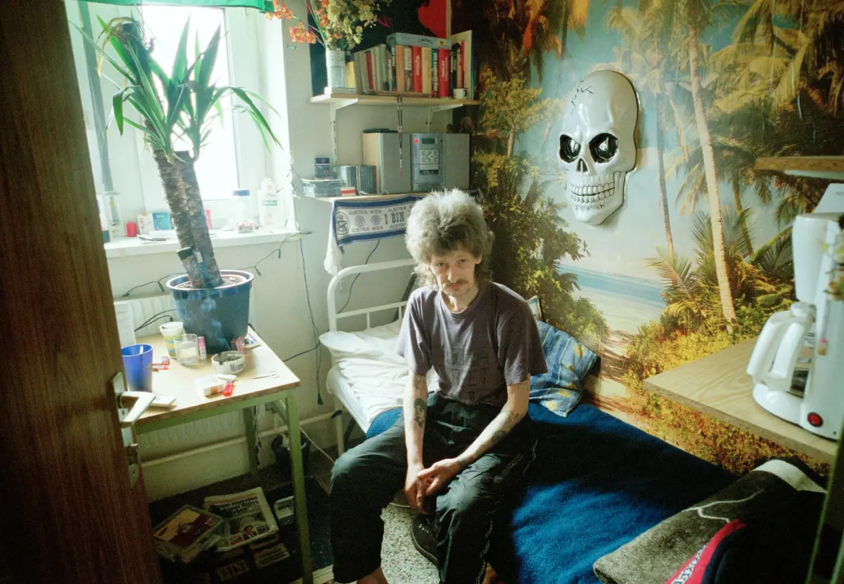 Person with curly hair and mustache sits on a bed in a small cluttered room with palm-tree mural and skull wall decor.