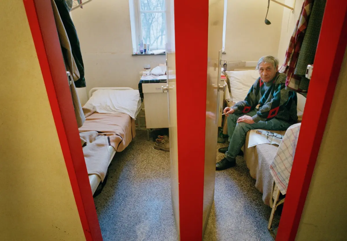 Elderly person seated on a bed in a small hospital room with two beds, red wardrobe doors, and a window in the background.