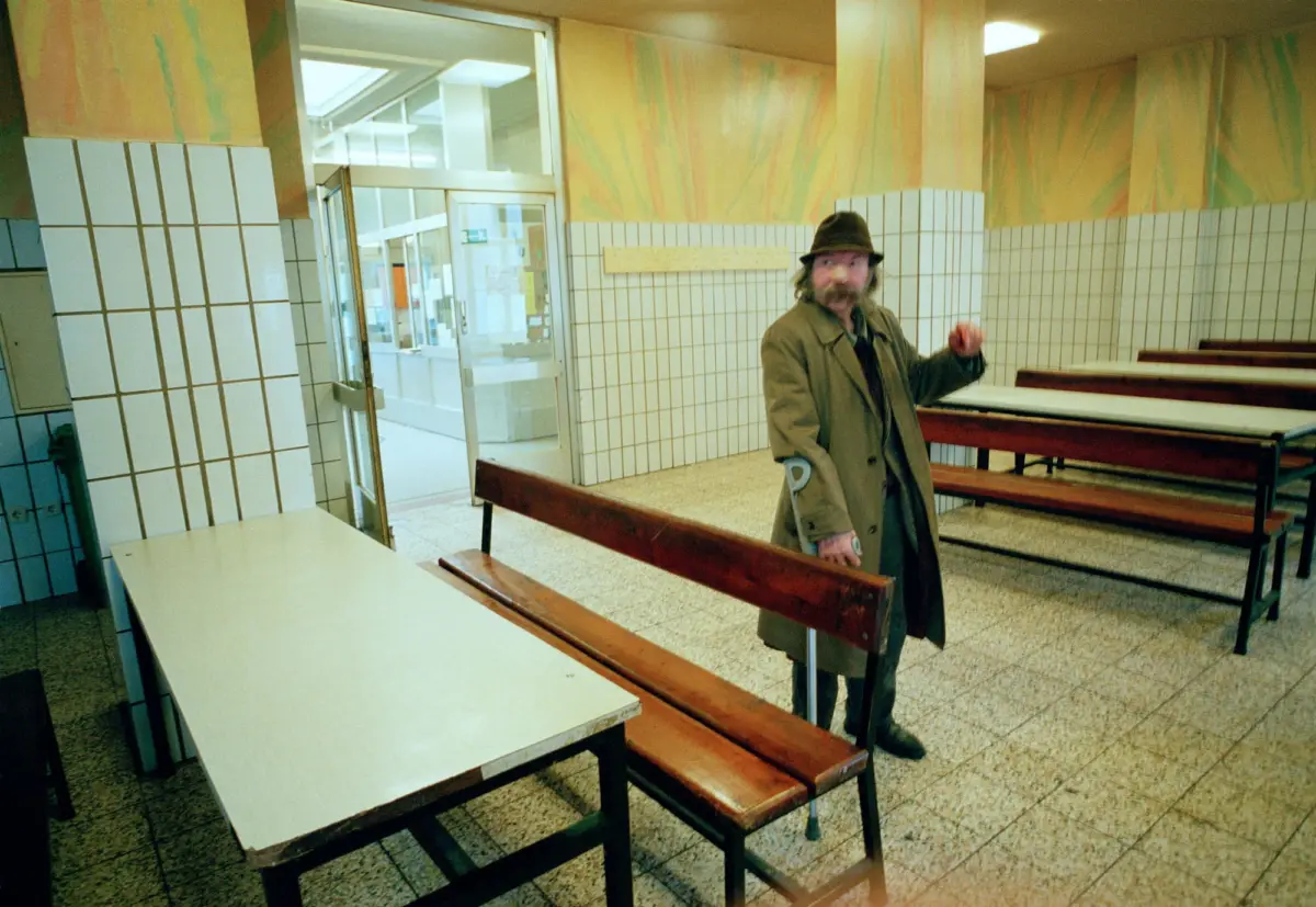 Person in a brown trench coat and hat stands beside wooden benches in a tiled, pastel-walled cafeteria.