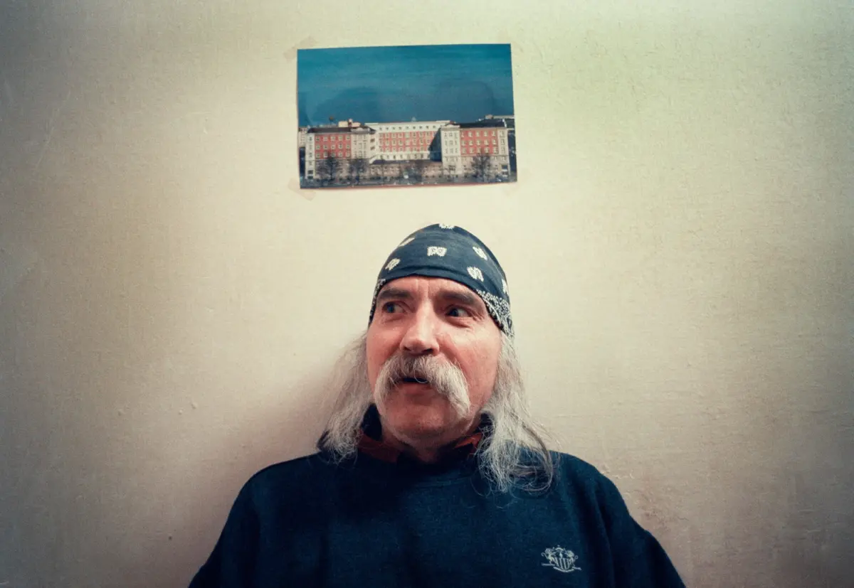 Older man with long white beard and bandana, looking up, against a beige wall with a photo of apartment buildings taped above.