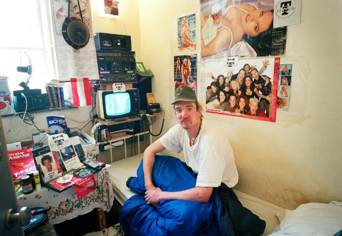 Person seated on bed with blue sleeping bag in a cluttered room with posters and vintage stereo equipment.