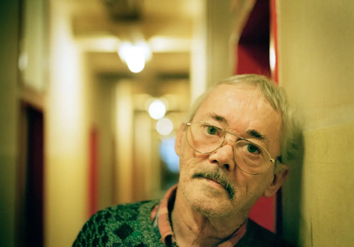 Older man with glasses leaning against a wall in a warm-lit indoor hallway, peering at the camera.