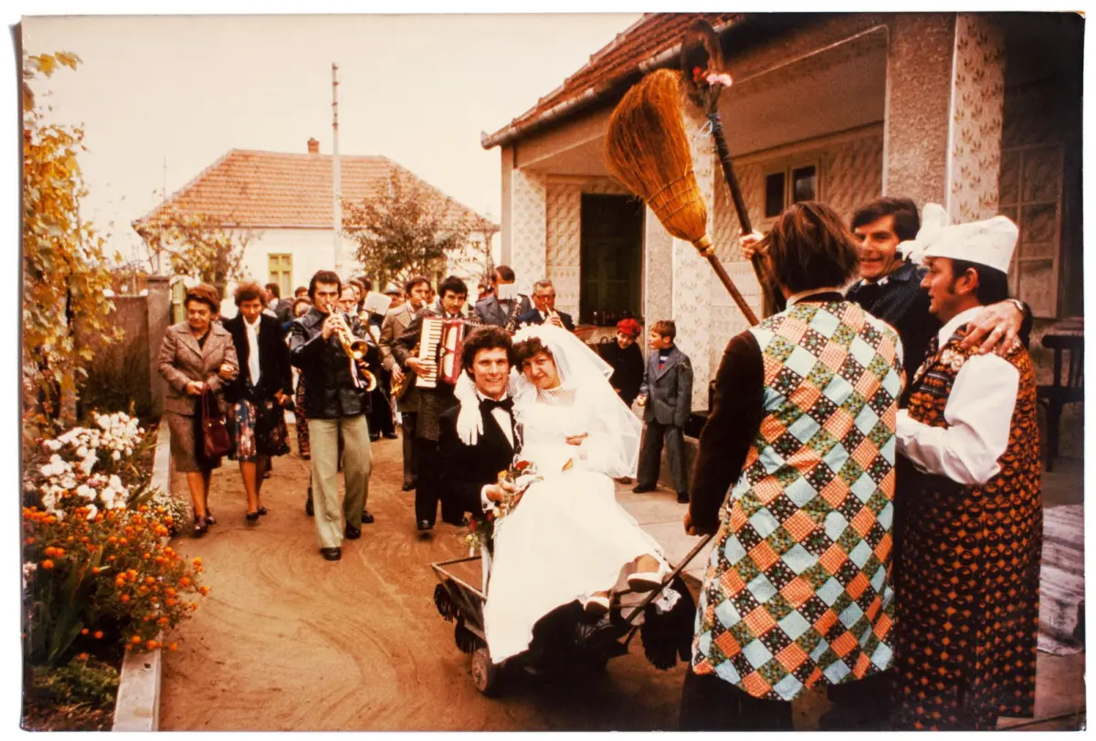 Bride and groom walk in wedding procession as musicians perform; vintage-clad onlookers line a village street.