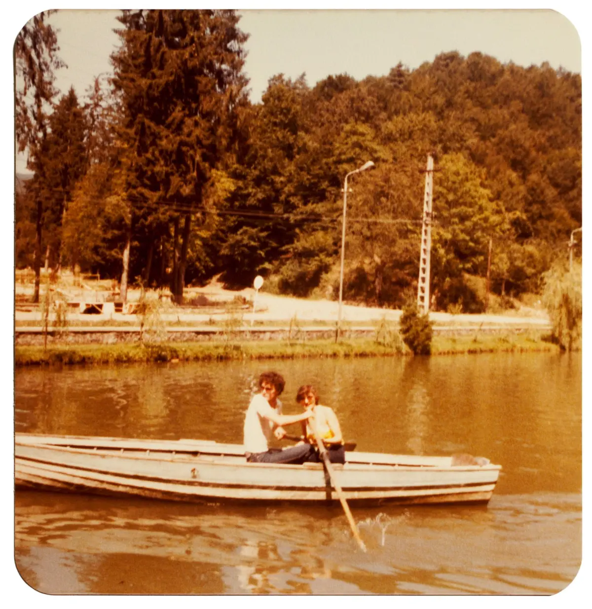 Two people sit in a weathered wooden rowboat on calm water by a tree-lined shore.