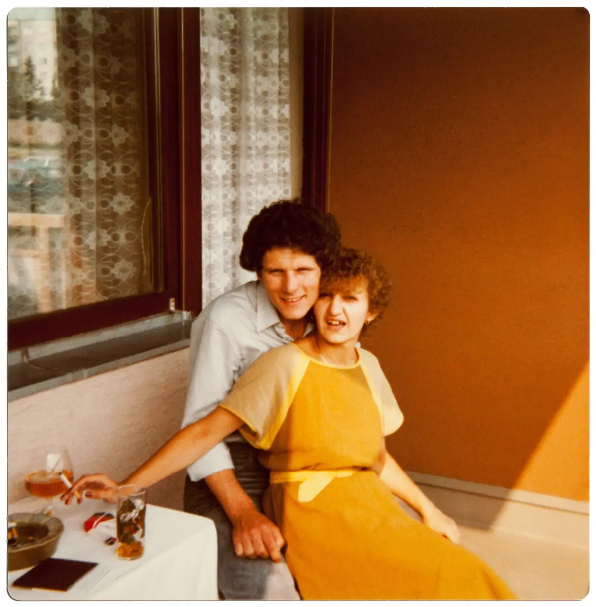 Two adults smiling for a photo on a balcony with an orange wall and lace-curtained windows; table with food and drink in foreground.