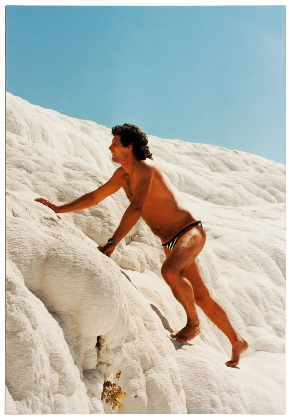 Shirtless adult wearing striped swim briefs climbs white textured rock against a blue sky