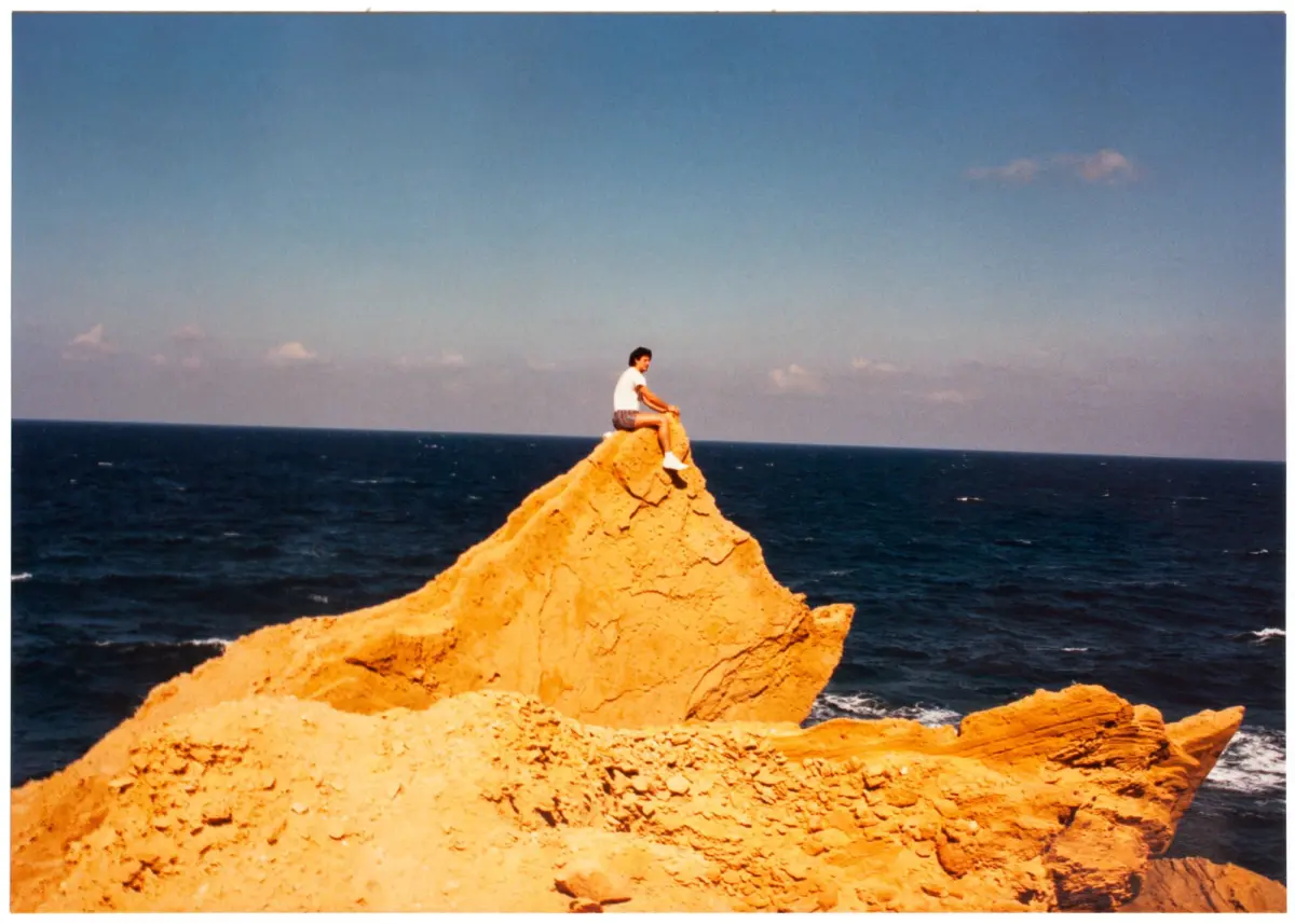 Person sitting on a jagged orange rock formation by the sea, gazing toward the water under a clear blue sky.