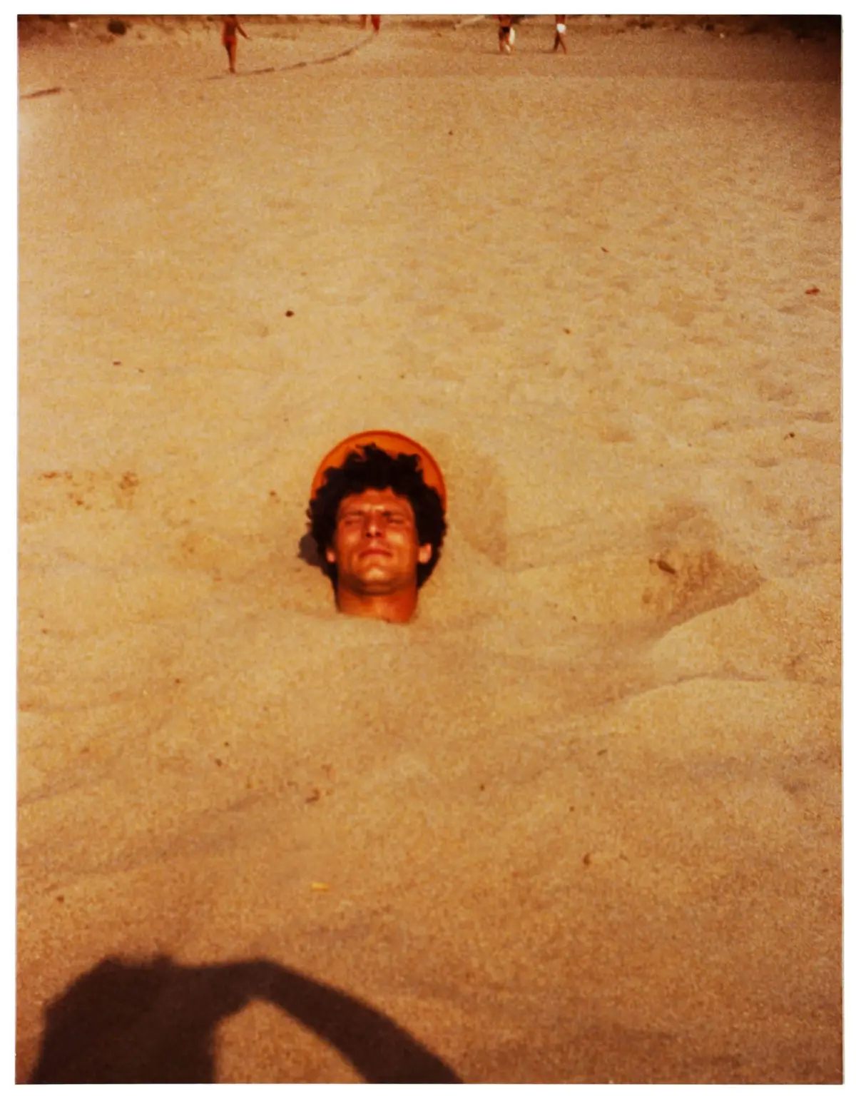 Person with orange hat partially buried in sand on a sunny beach; photographer's shadow visible in foreground.