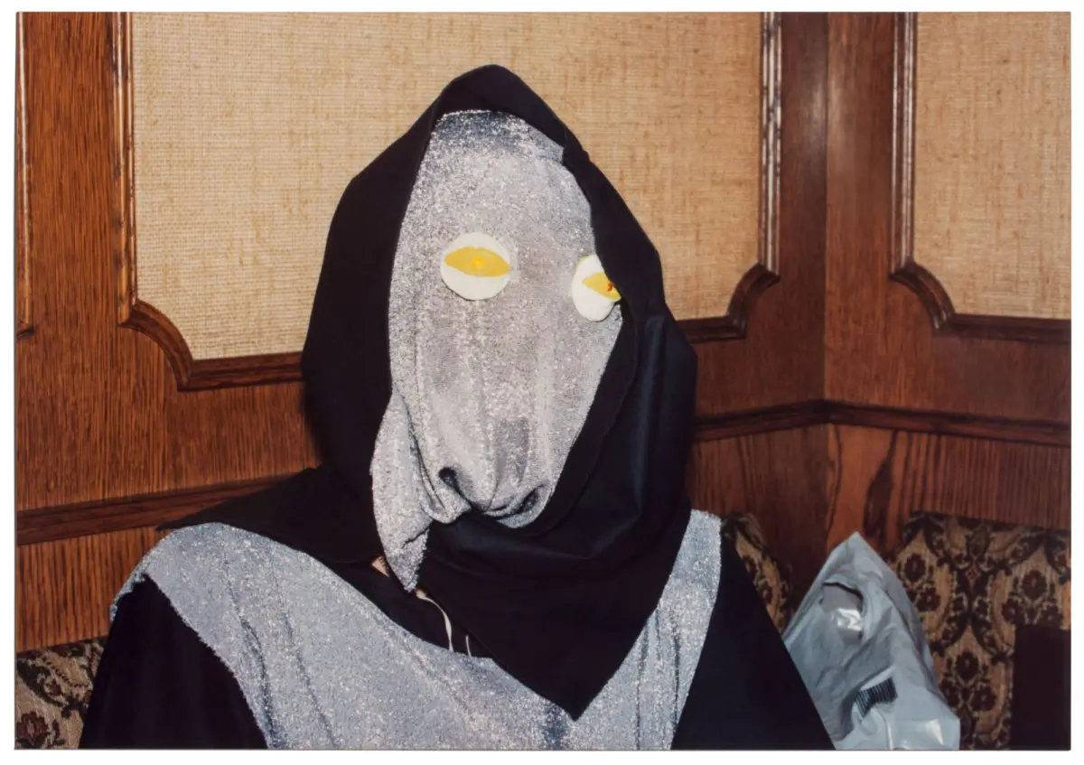 Person in glittery gray robe with black hood and yellow eye mask, seated on a patterned sofa in a wood-paneled room.