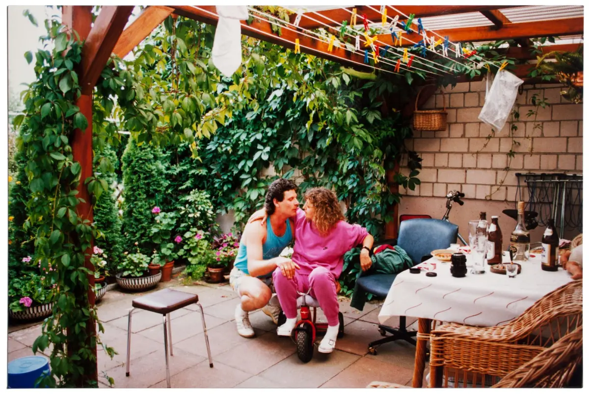 Couple cuddling on a small red tricycle in a vine-covered backyard patio under a wooden pergola.