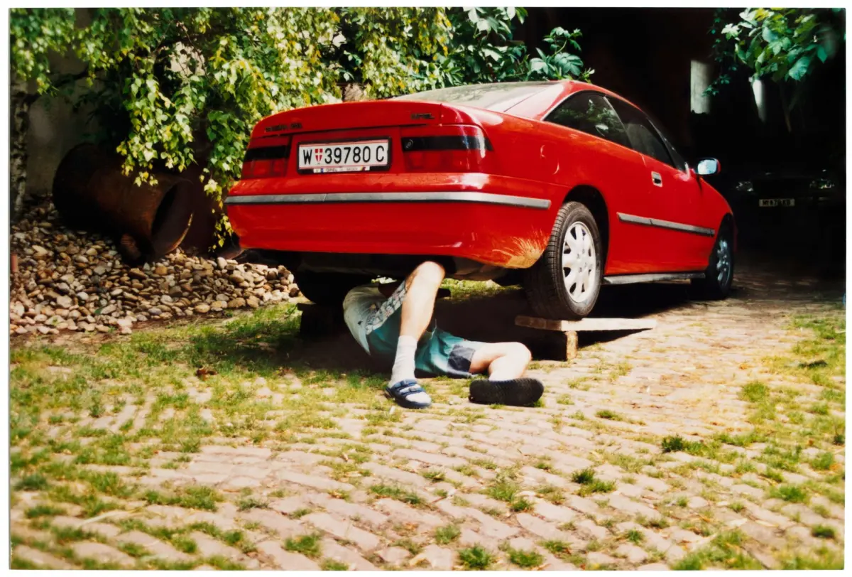 Red sports car raised on blocks; person lying under it, repairing undercarriage on a gravel driveway surrounded by greenery.