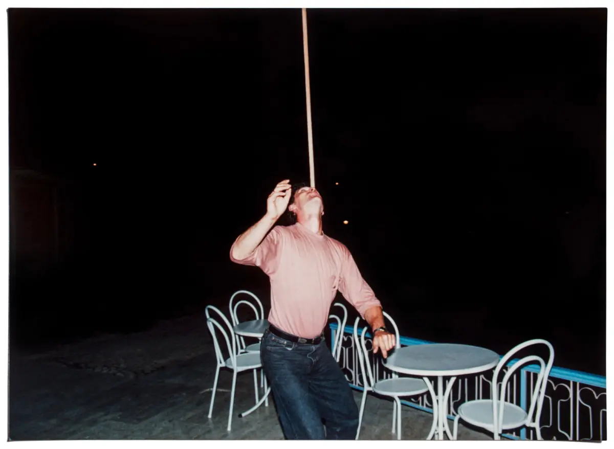 Nighttime outdoor cafe; person in pink shirt reaches toward a tall vertical pole, surrounded by white metal chairs around round tables.